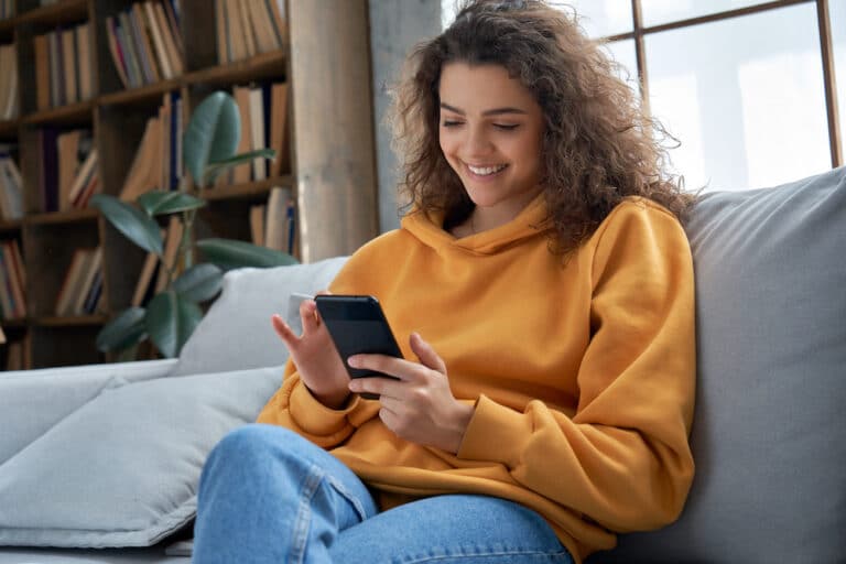 A young woman is sitting on a couch and using her phone to research fertility treatment options.