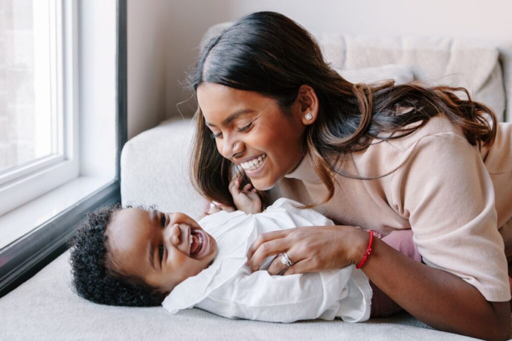 A woman smiles while holding her baby on a couch.